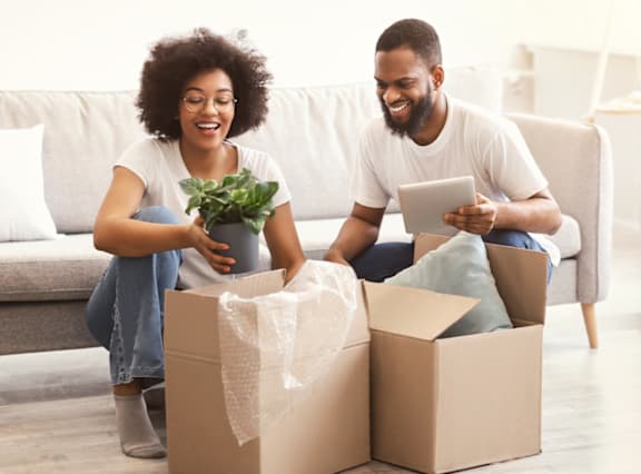 A man and woman are sitting on a couch with a potted plant and a tablet, surrounded by two cardboard boxes.