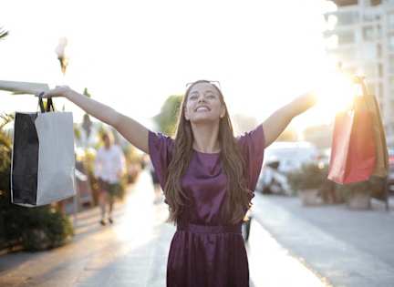 A woman in a purple dress is holding shopping bags and smiling.