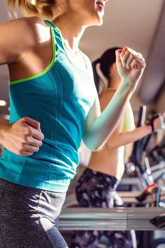 A woman in a blue tank top and grey leggings is running on a treadmill in a gym.