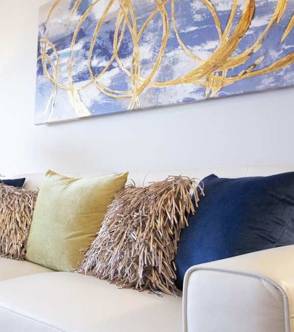 A white couch with blue and yellow pillows and a painting on the wall.