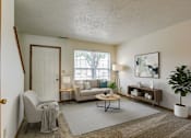 Thumbnail 29 of 31 - A living room with a grey sofa, a grey armchair, a wooden coffee table, a white side table, a white lamp, a white door, a white wall, a brown rug, a brown floor, a brown table, a