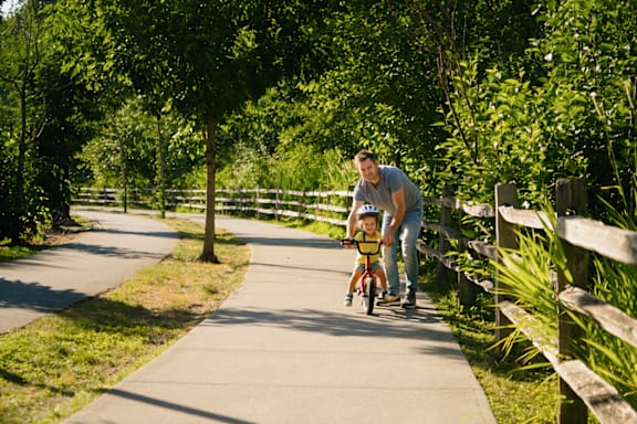 A man is pushing a child on a bike in a park.