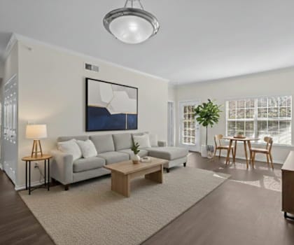 A living room with a grey couch, a coffee table, a lamp, a painting, a dining table with chairs, and a flat-screen TV mounted on the wall.