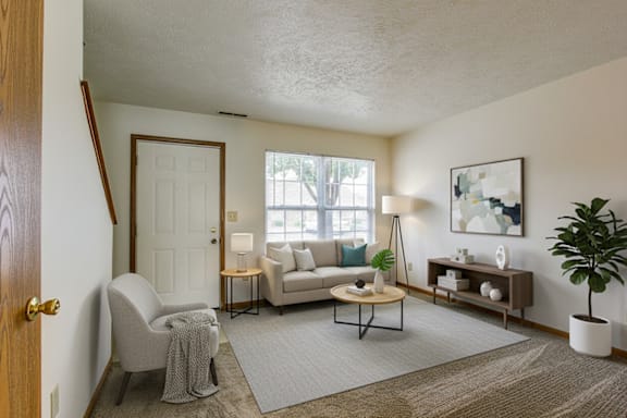 A living room with a grey sofa, a grey armchair, a wooden coffee table, a white side table, a white lamp, a white door, a white wall, a brown rug, a brown floor, a brown table, a