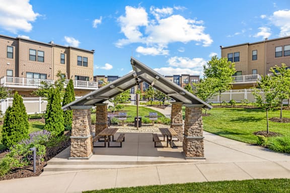 A pavilion with a stone pillar and a roof is surrounded by a grassy area and apartment buildings.