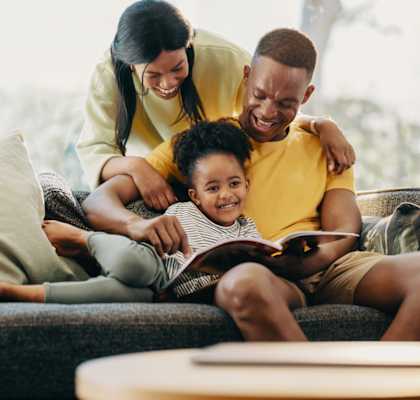 A family of three is sitting on a couch, with the child reading a book.