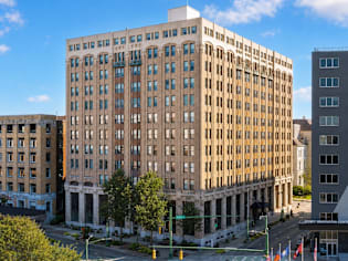 A large, beige building with many windows is in the foreground of a city street.