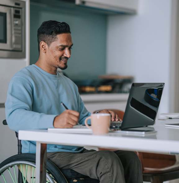 A man in a blue sweater is sitting at a table with a laptop and a cup of coffee.