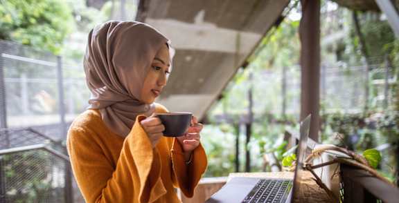 A woman in a yellow top and floral skirt is sitting at a wooden table with a laptop and a cup in front of her.