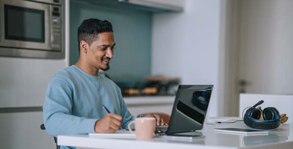 A man in a blue sweater is sitting at a table with a laptop and a cup of coffee.
