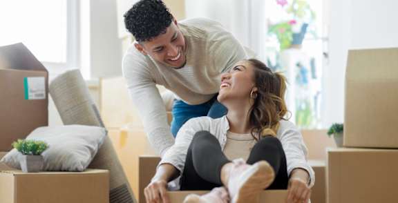 A man and woman are playing in a cardboard box in a room with other boxes and a couch.