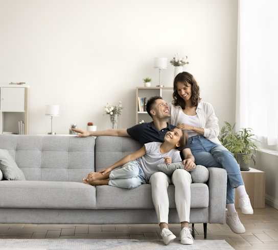 A woman and a child are sitting on a grey couch with a man lying on the floor behind them.