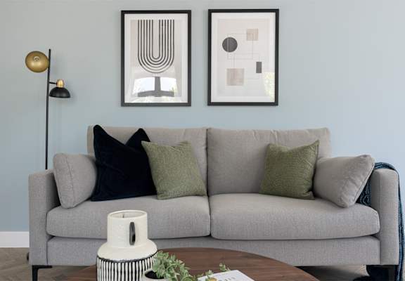 A grey couch with two green pillows and two framed artworks on the wall.