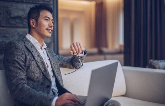 A man in a grey suit is sitting on a white couch using a laptop.
