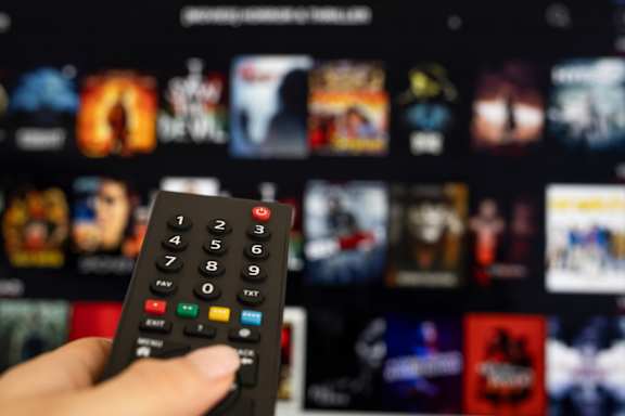 A hand is holding a remote control in front of a blurred background of a television screen displaying various shows.