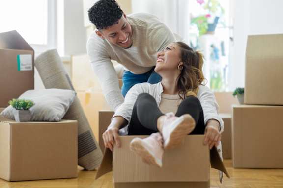 A man and woman are playing in a cardboard box in a room with other boxes and a couch.