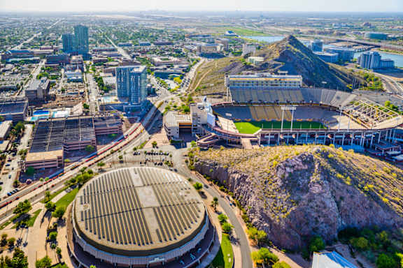 A large stadium with a grassy hill in front of it is surrounded by buildings.