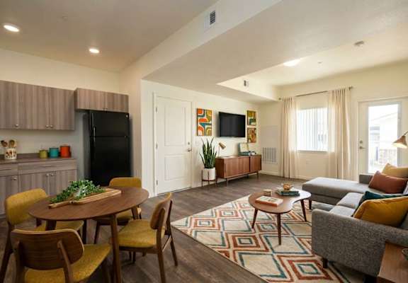 A living room with a grey couch, a wooden table and chairs, a black fridge, a flat screen TV, and a rug.
