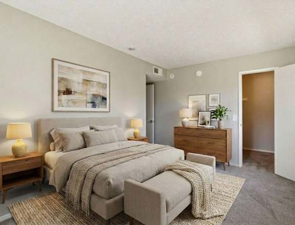 A bedroom with a large bed, a nightstand with a lamp, and a dresser with a plant on top.