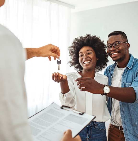 A person is handing over a key to a couple who are smiling and looking at a document.