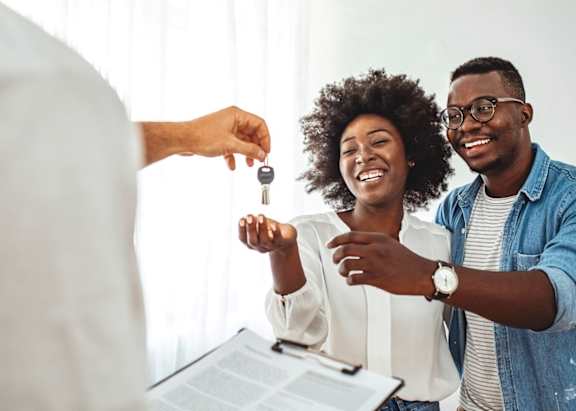 A person is handing over a key to a couple who are smiling and looking at a document.