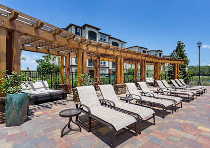 A row of lounge chairs are set up under a wooden pergola on a brick patio.