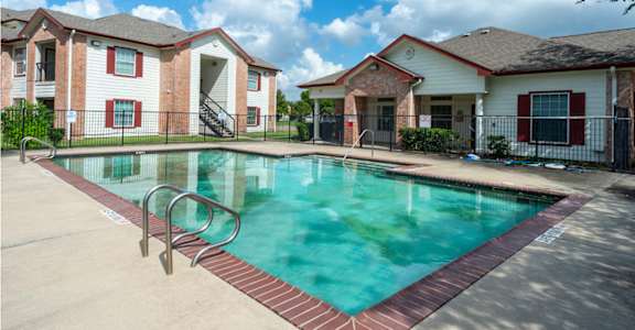 A swimming pool surrounded by a red brick border in front of apartment buildings.