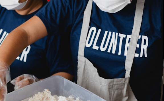 Two people wearing blue shirts with the word "VOLUNTEER" on them are handling food in a plastic container.