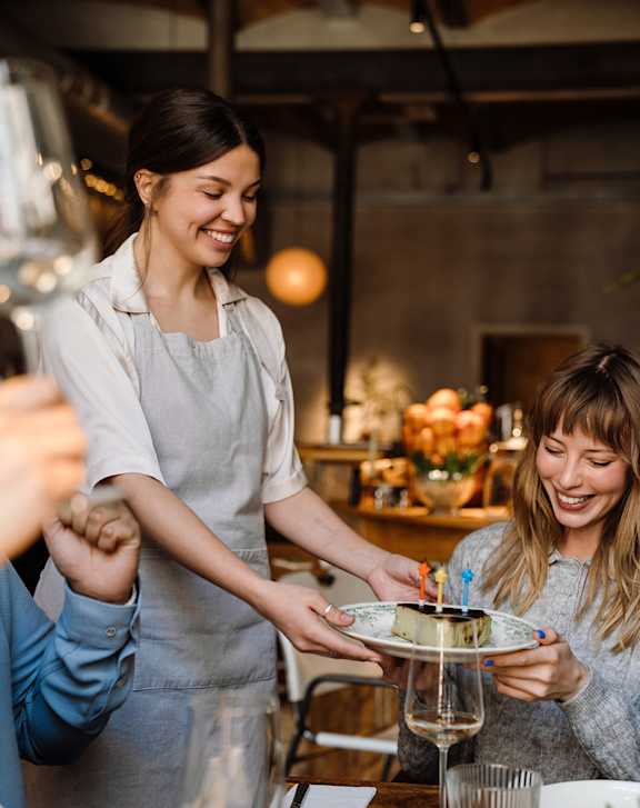 A woman in a grey apron is serving food to a woman in a blue shirt at a table.