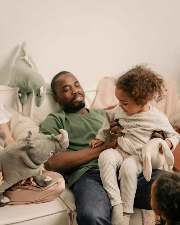A man is holding a child while sitting on a couch with stuffed animals around them at Fernlow Apartments, Houghton Regis