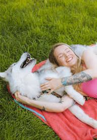 A woman is lying on the grass with her husky dog.