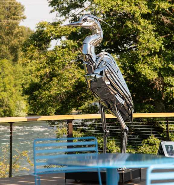 A metal sculpture of a bird is in front of a green tree.