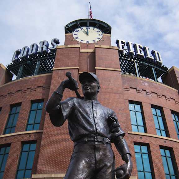 A statue of a baseball player stands in front of a brick building with a clock on top.