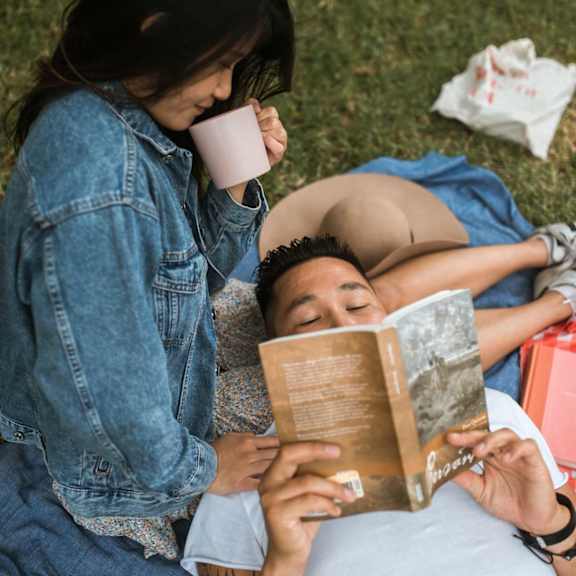A person is reading a book while another person is drinking from a cup.