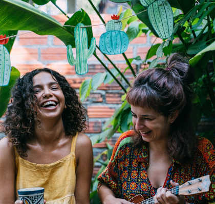 Two women sitting on a bench, one holding a cup and the other playing a ukulele.