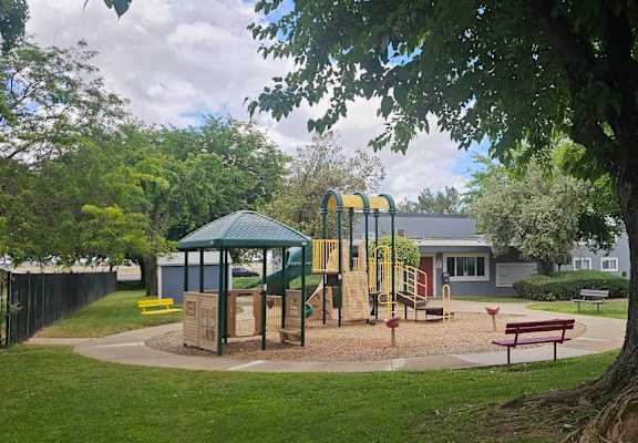 A playground with a green canopy and yellow slide is surrounded by a fence and a large tree.