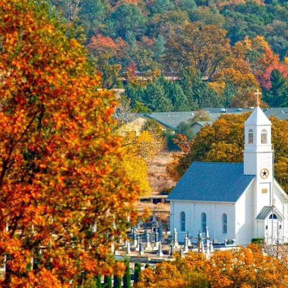 A white church with a bell tower is surrounded by trees with orange and yellow leaves.