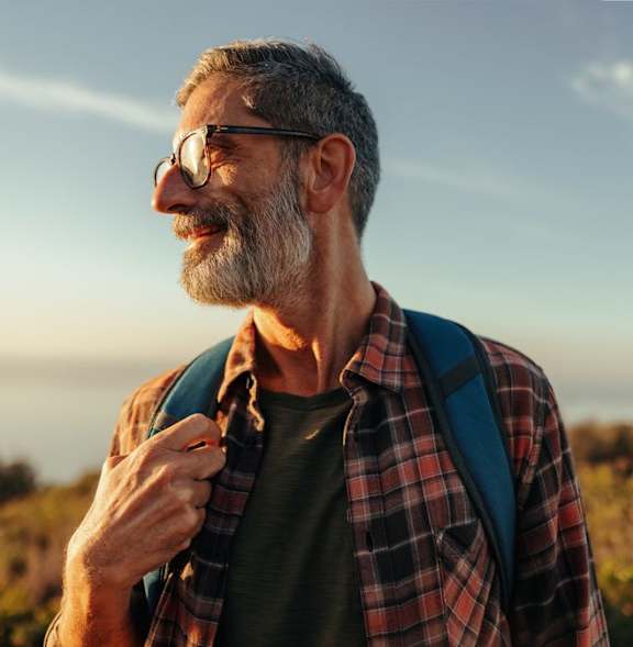 A man with a beard and glasses is standing outdoors wearing a plaid shirt and a backpack.