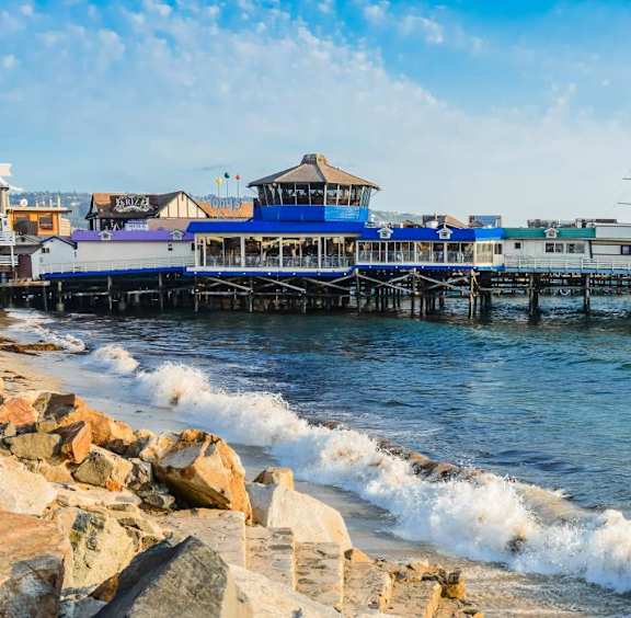 A pier with a building on it and waves crashing against the rocks.