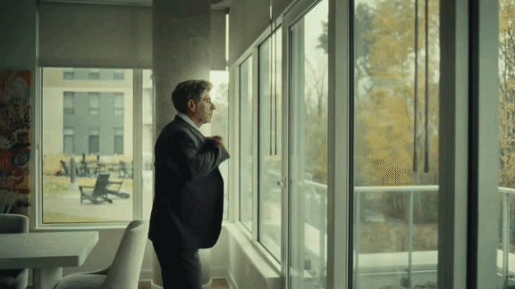 A man in a black suit stands by a window looking out at a tree-lined street.