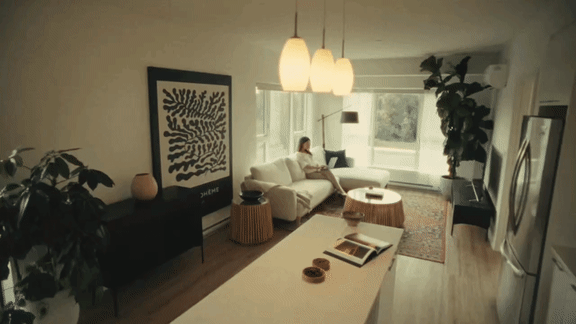 A woman is standing in a living room with a couch, a coffee table, and a large painting on the wall.