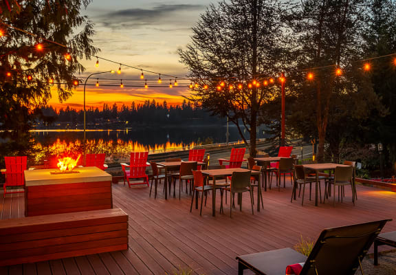 a deck with tables and chairs and a fire pit overlooking a lake at sunset