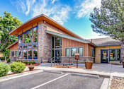 Thumbnail 1 of 26 - Aurora, CO Apartments Near Me - Copper Flats - Exterior View of Leasing Office with Parking Spaces, Seating Benches, Manicured Shrubs, Potted Plants, and Surrounding Trees.
