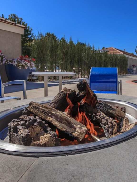 A fire pit with logs burning in the middle of a patio area with blue chairs and a pool in the background.