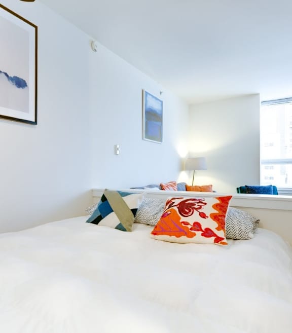 A bed with a white comforter and a blue pillow is in the foreground of a room with a framed picture of a blue sky with clouds on the wall.
