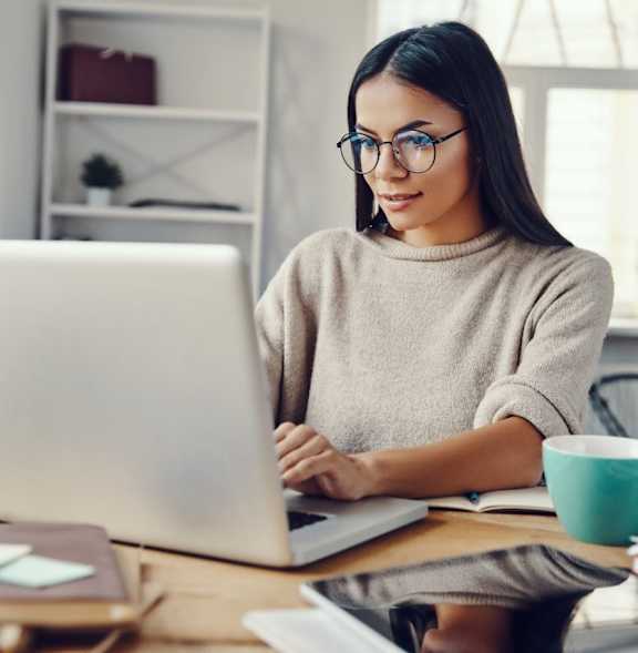 A woman is working on her laptop at a desk with various items scattered around.