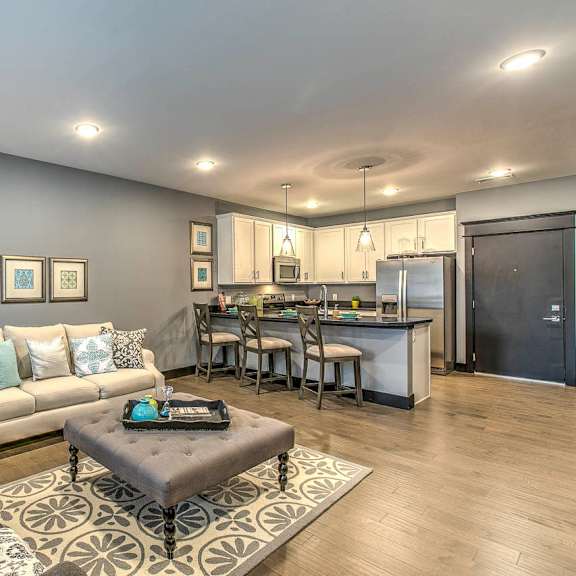 Modern apartment kitchen with white cabinetry, stainless steel appliances, and a dark countertop island with three bar stools and pendant lighting above.