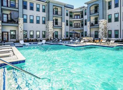 A swimming pool in front of a building with a deck and lounge chairs.