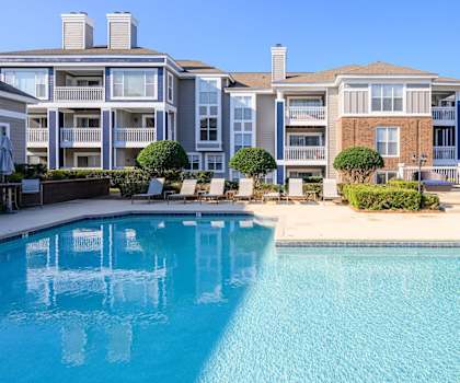 The large, turquoise outdoor pool with pool chairs, tables, and chairs with the apartments in the background at Retreat at McAlpine Creek Apartment Homes. 