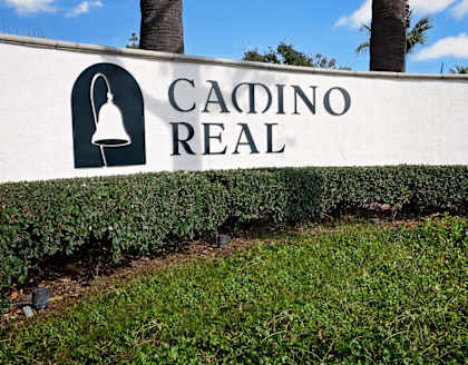 A white sign with the words "CADINO REAL" and a bell icon in front of a hedge and palm trees.